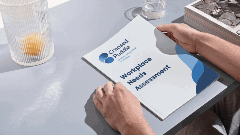 Person holding a booklet titled "Workplace Needs Assessment" with the Creased Puddle logo and tagline "Understanding Minds of All Kinds" on the cover, placed on a grey desk beside a glass of water, a ridged glass with an orange inside, and a book with glasses on top.