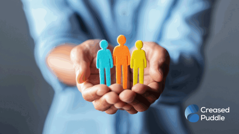 Person holding three colourful cut-out human figures in their hands, symbolising diversity and inclusion, with the Creased Puddle logo in the bottom right corner.