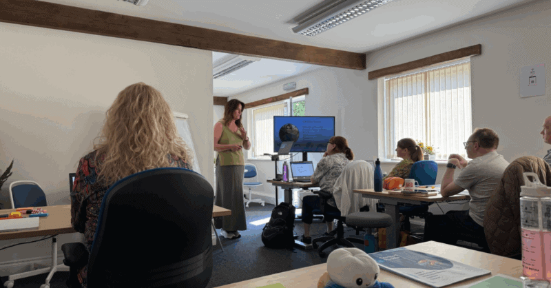 A woman stands at the front of a well-lit training room delivering a presentation. A slide on the screen shows the name "Caroline Turner" with her photo and title. Several attendees sit at desks facing her, engaged and taking notes. The setting is casual and comfortable, with natural light coming through the windows.