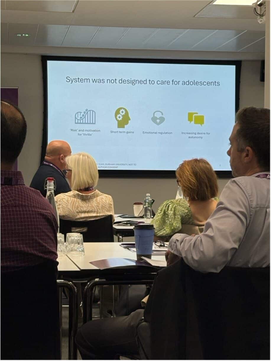 Audience members seated at a conference watching a presentation slide that reads “System was not designed to care for adolescents.” The slide includes four icons with labels: “Risk and motivation for ‘thrills’,” “Short term gains,” “Emotional regulation,” and “Increasing desire for autonomy.”