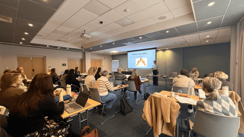 Professionals attending a neurodiversity workplace training session, with a presenter at the front of the room and a projected slide reading "Before we start..."