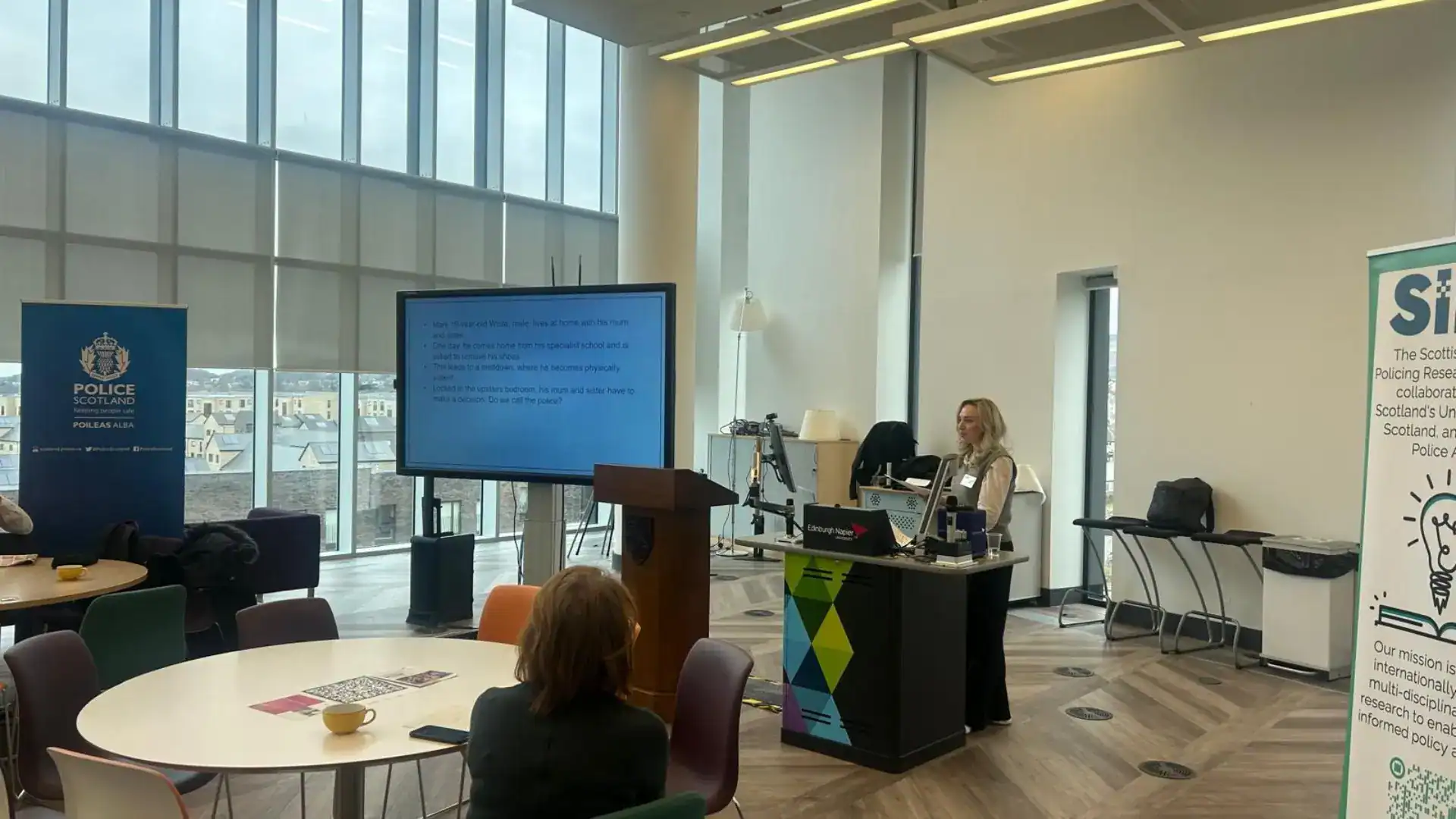 A speaker delivers a presentation in a modern conference room with large floor-to-ceiling windows. Attendees are seated at round tables while a presentation slide is displayed on a large screen. Banners for Police Scotland and the Scottish Institute for Policing Research are visible on either side of the room.